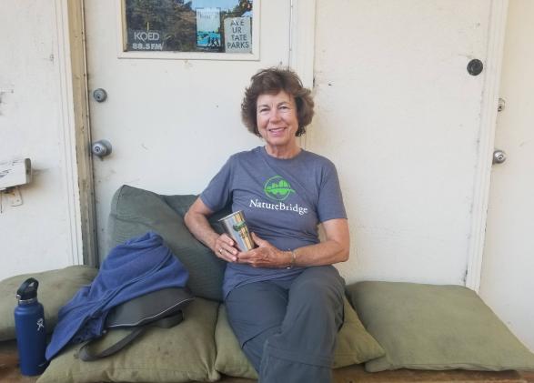 NatureBridge educator Ingrid Apter sitting against a white wall with a cup in hand sitting on green cushions